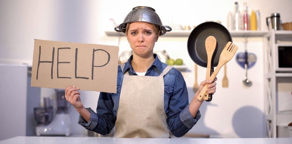 Fundraising Lunch - A white female presenting person in a jean shirt and canvas apron with a metal strainer on their head like a hat while standing in a kitchen. In one hand they are holding a cardboard sign with the word help written on it, and in the other hand are wooden utensils and a cast iron pan.