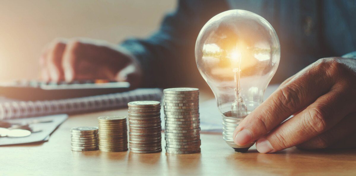 diversity and inclusion initiatives - A close-up of a person of color's hands at a table. The hand in the foreground is holding a lit lightbulb between their fingers and resting the base on the table. There are four stacks of coins of increasing height leading up to the lightbulb. In the background you see the person's other hand typing into a calculator that is sitting on top of a folder.