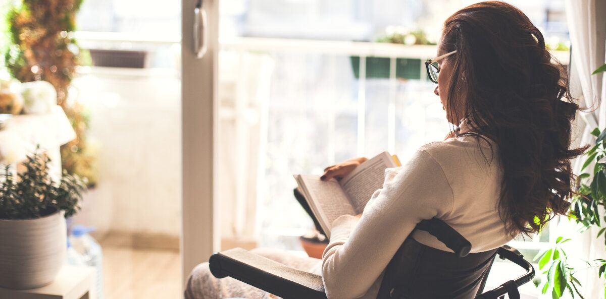nonprofit books - An over the shoulder view of a brown skinned female presenting person in a wheelchair reading a book in a room with lots of windows and plants