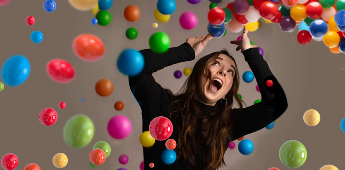 Image of a woman in a black long sleeve holding her head above her arms with a shocked look on her face as multi-colored balls fall down on her. Suggests the need for better time management.