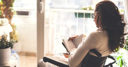 nonprofit books - An over the shoulder view of a brown skinned female presenting person in a wheelchair reading a book in a room with lots of windows and plants