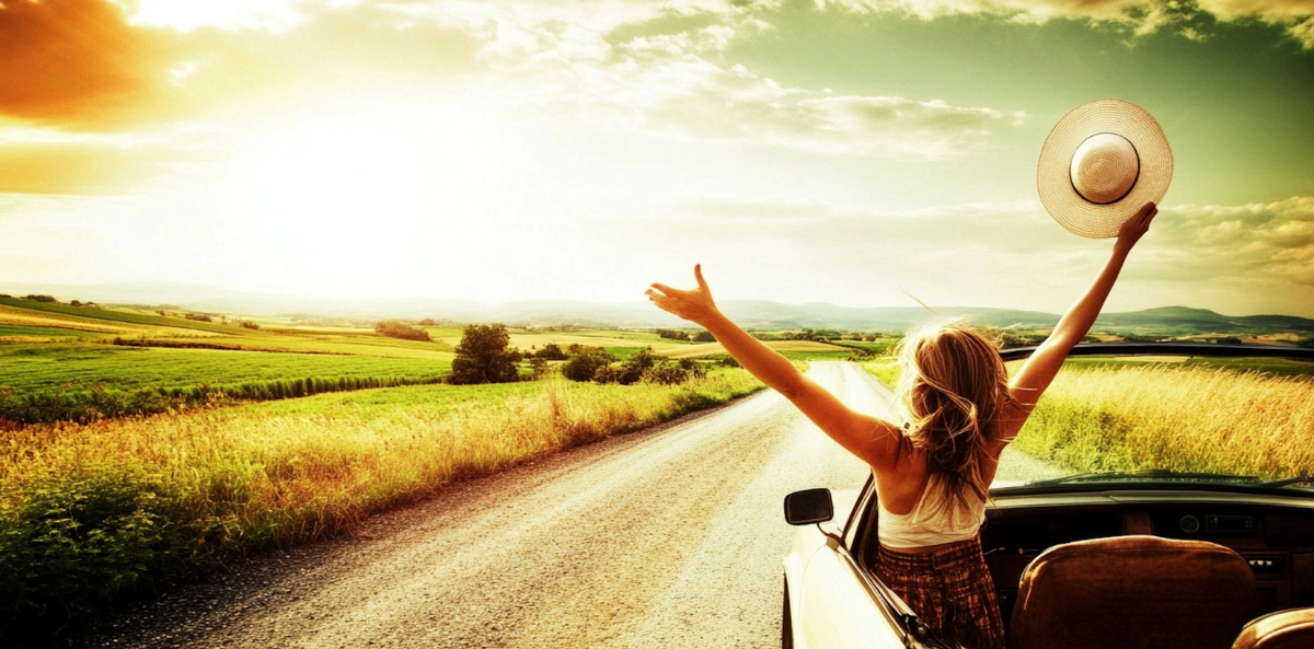 A woman with open arms is in the driver's seat of a car with a big open sky in front of her.