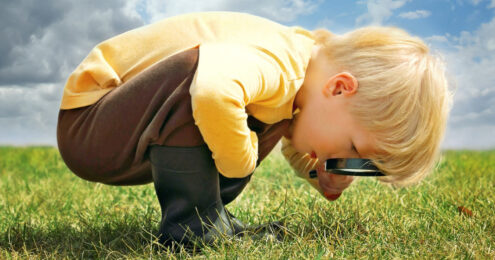 micromanage - A young boy with blonde hair wearing a long sleeve yellow shirt and brown pants tucked into black rain boots is crouched and bent over with a magnifying glass pressed to his eyes intently looking at something in the grass